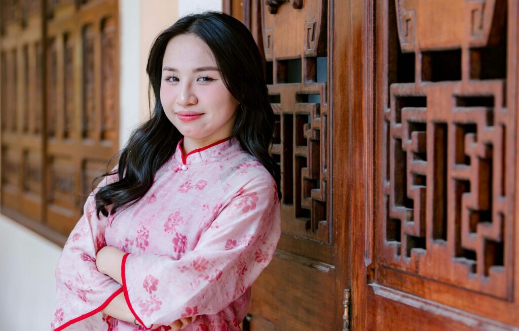 Smiling woman wearing a pink cheongsam stands by wooden doors.